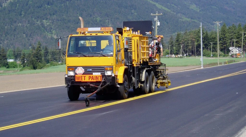 A British Columbia highway maintenance truck spraying white lane markings on the asphalt, flanked by orange traffic cones and a flashing arrow board.