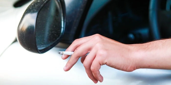 Close-up shot of a driver's hand holding a lit cigarette out of an open car window on a highway shoulder.