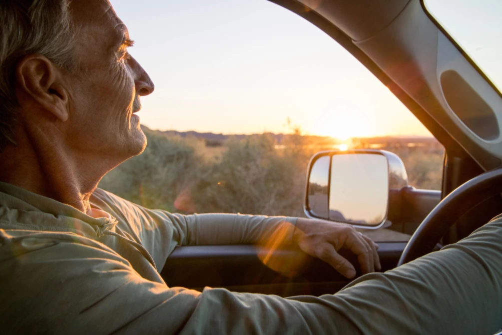 An older male seen from the passenger seat driving a vehicle into the sunset